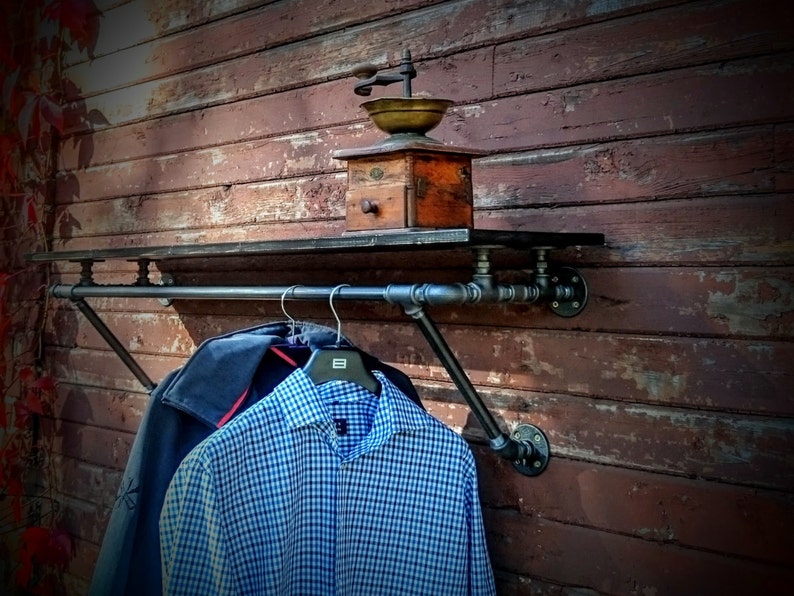 May include: A black metal pipe shelf with a black metal pipe rod attached to the bottom. The shelf is mounted to a wooden wall and has a vintage coffee grinder on top. Two shirts are hanging on the rod.