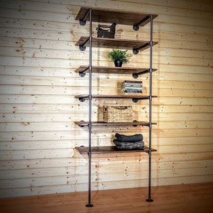 May include: A six-shelf industrial pipe shelving unit with wooden shelves. The shelves are made of dark brown wood and the pipes are black metal. The unit is standing against a wood-paneled wall. There are items on the shelves, including a small plant, books, a woven basket, and folded clothing.