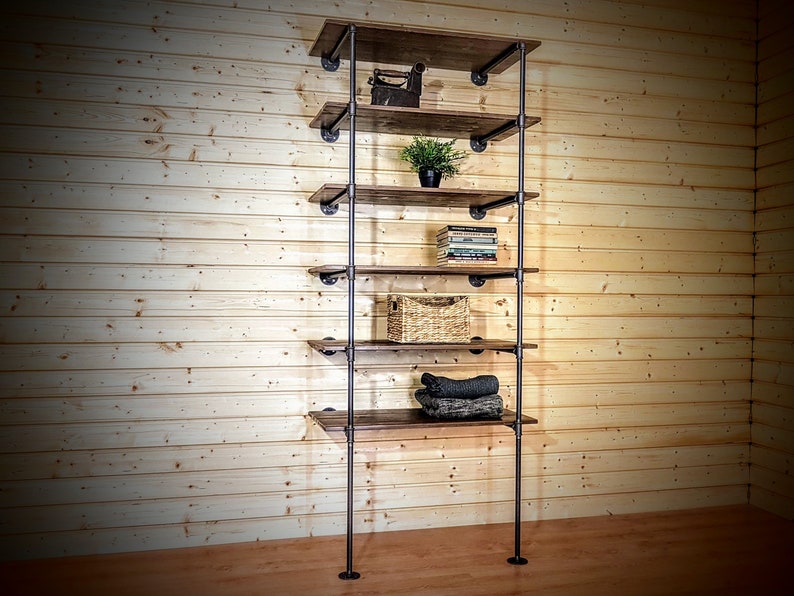 May include: A six-shelf industrial pipe shelving unit with wooden shelves. The shelves are made of dark brown wood and the pipes are black metal. The unit is standing against a wood-paneled wall. There are items on the shelves, including a small plant, books, a woven basket, and folded clothing.