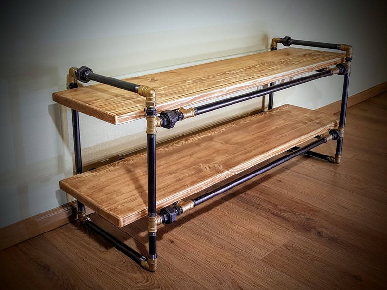May include: A two-tiered shelf made of black metal pipes and wooden planks. The shelf has gold-colored fittings and is sitting on a wooden floor.