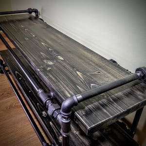 May include: A black metal and wood shelf with two tiers. The shelf is made of industrial pipes and has a dark wood finish.