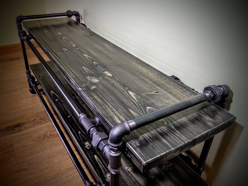 May include: A black metal and wood shelf with two tiers. The shelf is made of industrial pipes and has a dark wood finish.