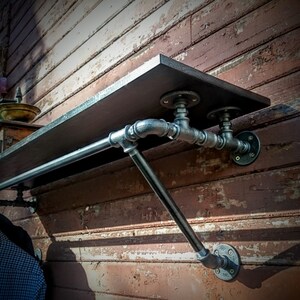 May include: A black metal pipe shelf with a wooden top and a metal rod for hanging clothes. The shelf is mounted on a brick wall.