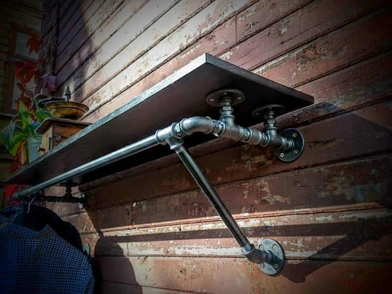 May include: A black metal pipe shelf with a wooden top and a metal rod for hanging clothes. The shelf is mounted on a brick wall.