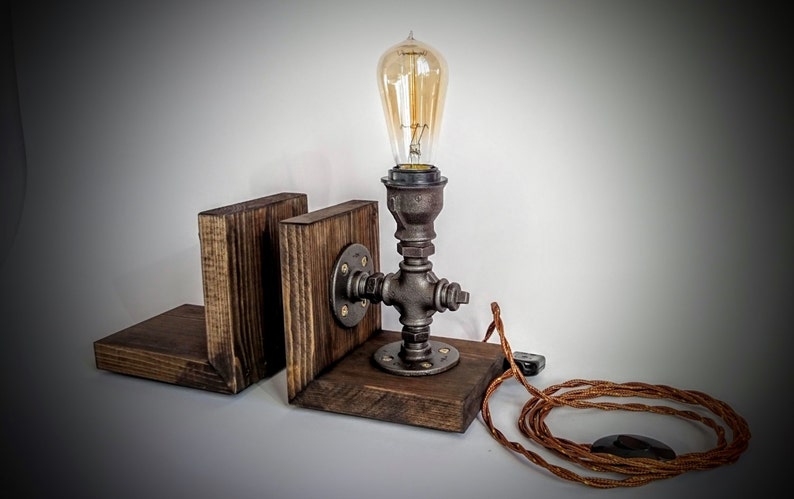 May include: A rustic wooden bookend lamp with a black metal pipe base and a vintage Edison bulb. The lamp has a brown cord and a black plug.