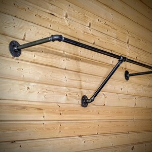 Puede incluir: Perchero de pared de estilo industrial hecho de tuber&iacute;as y accesorios de metal negro. El perchero tiene una barra horizontal para colgar ropa, soportada por tuber&iacute;as y bridas anguladas, montado en una pared de madera clara.