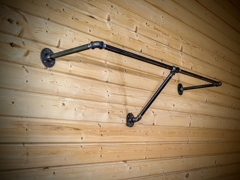 Puede incluir: Perchero de pared de estilo industrial hecho de tuber&iacute;as y accesorios de metal negro. El perchero tiene una barra horizontal para colgar ropa, soportada por tuber&iacute;as y bridas anguladas, montado en una pared de madera clara.