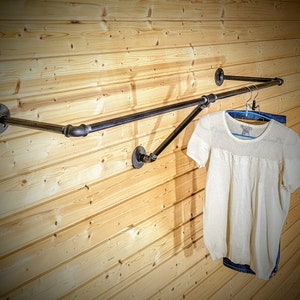 Puede incluir: Perchero de estilo industrial hecho de tuber&iacute;as de metal negro, montado en una pared de tablones de madera clara. Una camisa blanca de manga corta y unos vaqueros azules cuelgan del perchero. El perchero tiene una barra horizontal y un soporte diagonal.