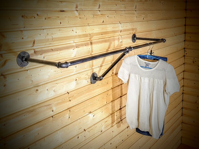 Puede incluir: Perchero de estilo industrial hecho de tuber&iacute;as de metal negro, montado en una pared de tablones de madera clara. Una camisa blanca de manga corta y unos vaqueros azules cuelgan del perchero. El perchero tiene una barra horizontal y un soporte diagonal.