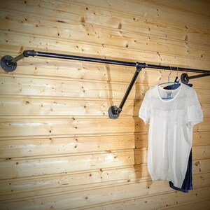 Puede incluir: Un perchero de ropa de tuber&iacute;a industrial negra montado en una pared de paneles de madera clara. Una camisa blanca de manga corta y unos vaqueros azules cuelgan del perchero. El perchero est&aacute; hecho de tuber&iacute;as y accesorios negros.