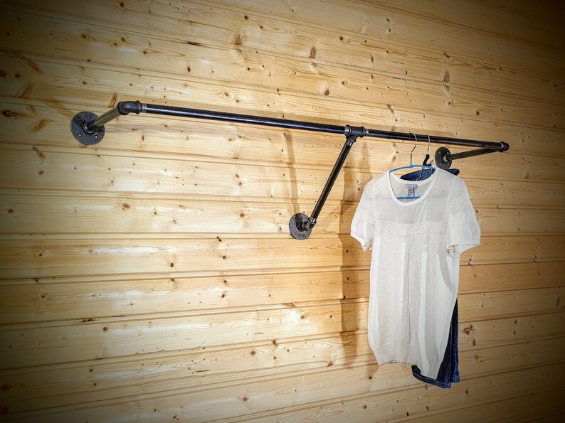 Puede incluir: Un perchero de ropa de tuber&iacute;a industrial negra montado en una pared de paneles de madera clara. Una camisa blanca de manga corta y unos vaqueros azules cuelgan del perchero. El perchero est&aacute; hecho de tuber&iacute;as y accesorios negros.