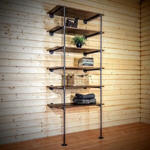 May include: A six-shelf industrial pipe shelving unit with brown wooden shelves. The shelves are filled with various items, including a plant, books, a basket, and folded towels.