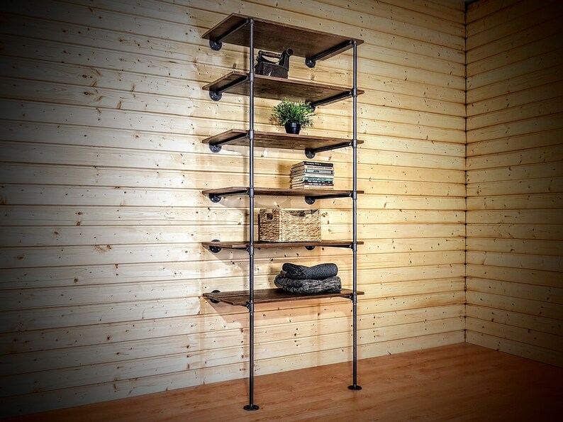 May include: A six-shelf industrial pipe shelving unit with brown wooden shelves. The shelves are filled with various items, including a plant, books, a basket, and folded towels.