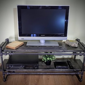 May include: A black metal TV stand with two shelves. The stand has a dark wood top and bottom shelf. A flat screen TV, a book, a plant, and a DVD player are on the stand.