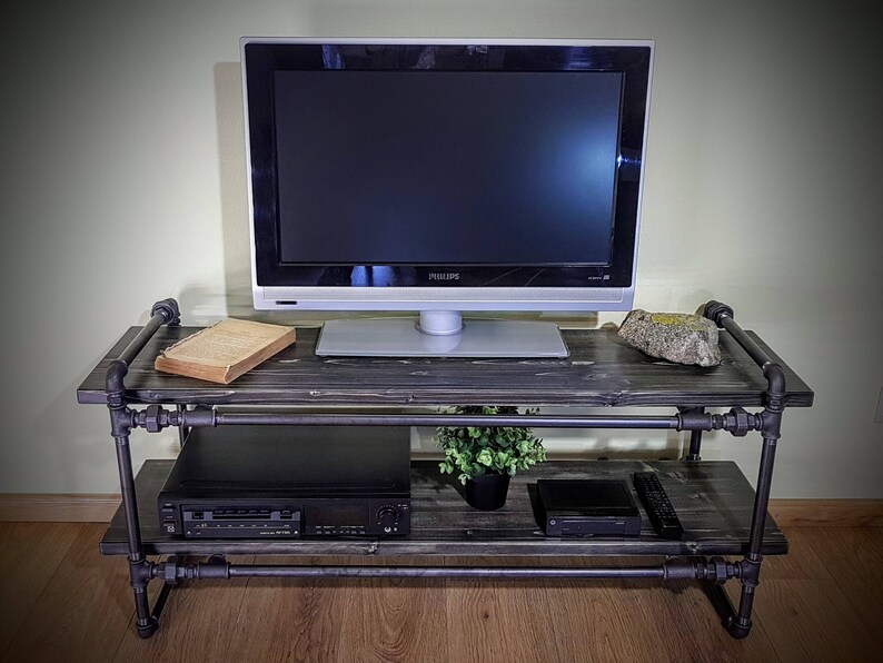 May include: A black metal TV stand with two shelves. The stand has a dark wood top and bottom shelf. A flat screen TV, a book, a plant, and a DVD player are on the stand.