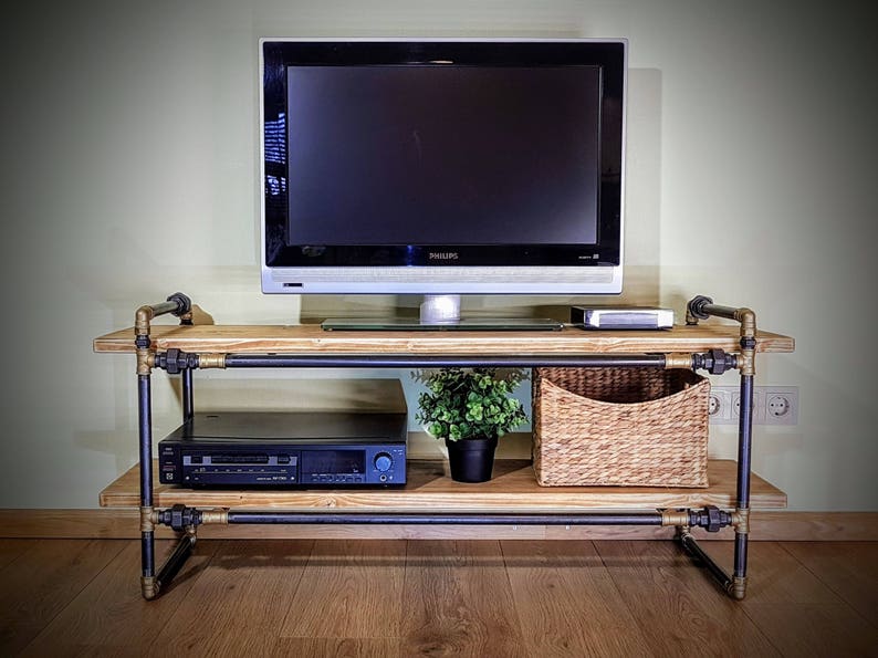 May include: A black metal TV stand with two shelves made of wood. The stand has gold-colored fittings and is holding a flat-screen TV, a stereo system, a potted plant, and a wicker basket.