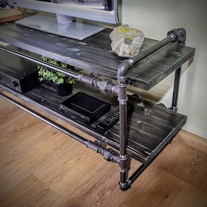 May include: Black metal pipe TV stand with two shelves. The stand has a black wood top and a black wood bottom shelf. There is a TV, a DVD player, and a plant on the shelves.