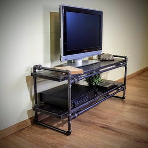 May include: A black metal TV stand with two shelves. The stand has a rustic industrial design and is made of pipes and wood. A flat screen TV is sitting on the top shelf.