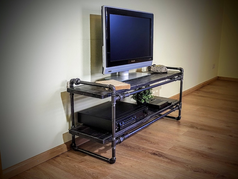 May include: A black metal TV stand with two shelves. The stand has a rustic industrial design and is made of pipes and wood. A flat screen TV is sitting on the top shelf.