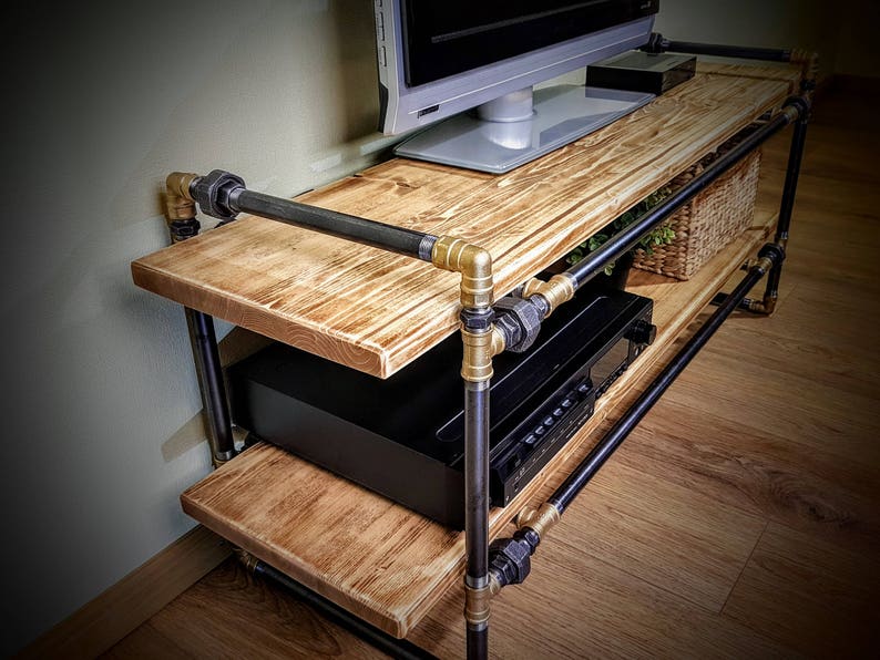 May include: A two-tiered industrial style TV stand with black metal pipes and wooden shelves. The stand has a gold finish on the pipe fittings and is holding a flat screen TV, a black stereo receiver, and a wicker basket.