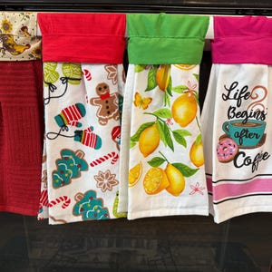 May include: Four decorative kitchen towels hanging on an oven handle. The towels feature various designs, including a rooster, Christmas motifs, lemons, and a coffee-themed towel with the text "Life Begins After Coffee."