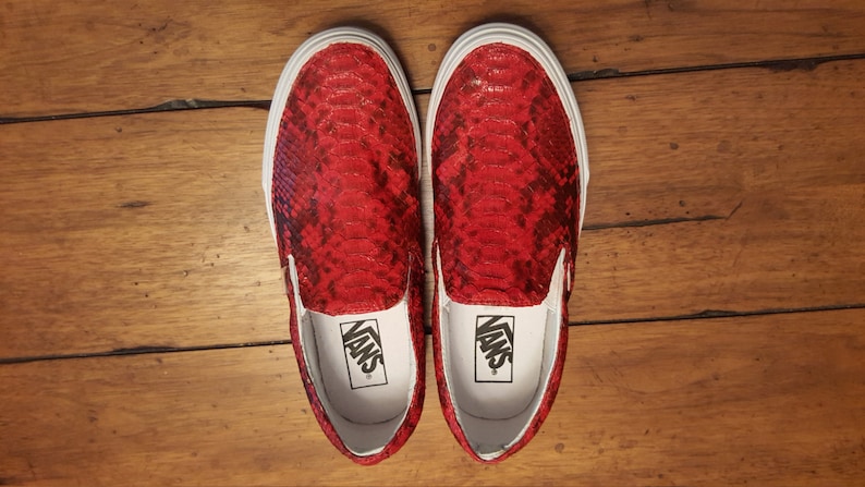 May include: A pair of red snakeskin Vans slip-on shoes with white laces and white soles. The shoes are on a wooden surface.