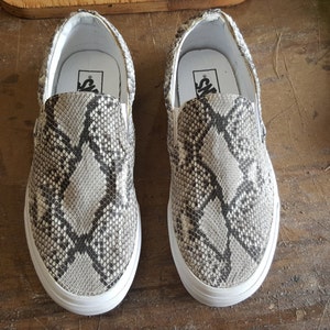 May include: A pair of white slip-on shoes with a snake skin pattern. The shoes have a black Vans logo on the back.