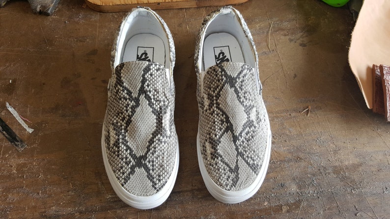 May include: A pair of white slip-on shoes with a snake skin pattern. The shoes have a black Vans logo on the back.
