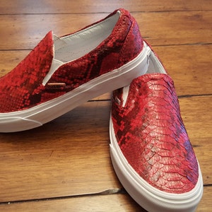 May include: A pair of red snakeskin Vans slip-on shoes with white soles.