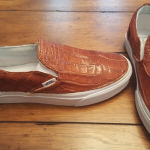 May include: A pair of brown leather slip-on Vans shoes with a crocodile pattern. The shoes have white soles and laces.