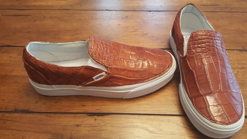 May include: A pair of brown leather slip-on Vans shoes with a crocodile pattern. The shoes have white soles and laces.