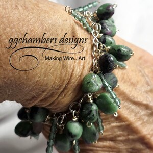 May include: A handmade bracelet with green and black gemstone beads and silver-colored chain links. The bracelet features the text "ggchambers designs Making Wire...Art". The beads are various sizes and shapes.