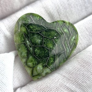 May include: A heart-shaped pendant with a green, marbled appearance, encased in a silver-colored wire frame. The pendant is set against a white background, highlighting its unique design and color variations. The piece has a polished, reflective surface.