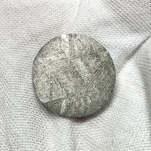 May include: A circular metallic object with a silver-toned surface featuring a distinct cross-hatched pattern of intersecting lines. The texture appears etched or naturally weathered, creating a complex geometric grid across the flat, round face.