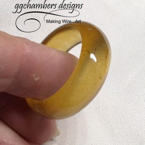 May include: A chunky, amber-toned ring with a smooth, polished finish. The ring is held between fingers, highlighting its rounded form and translucent appearance. The ring is a piece of jewellery, and the text "ggchambers designs Making Wire...Art" is in the background.