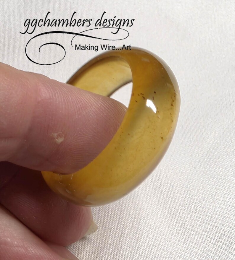 May include: A chunky, amber-toned ring with a smooth, polished finish. The ring is held between fingers, highlighting its rounded form and translucent appearance. The ring is a piece of jewellery, and the text "ggchambers designs Making Wire...Art" is in the background.