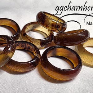 May include: A selection of amber and brown resin rings. The rings have varying patterns and colours, some with a marbled effect. The image includes the text "ggchambers designs Making Wire...Art".