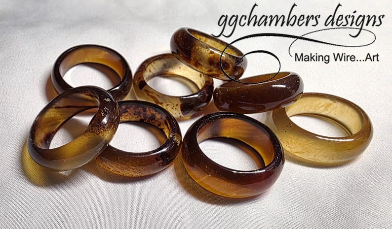 May include: A selection of amber and brown resin rings. The rings have varying patterns and colours, some with a marbled effect. The image includes the text "ggchambers designs Making Wire...Art".