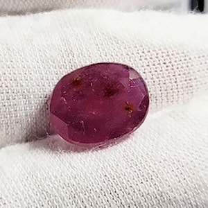 May include: A deep pink oval-shaped gemstone with a faceted cut. The gemstone has a slight reddish-brown hue and is set against a white background.