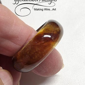 May include: An amber-coloured ring with a glossy finish. The ring has a unique, organic design with visible internal patterns. The text "ggchambers designs Making Wire...Art" is visible in the background.