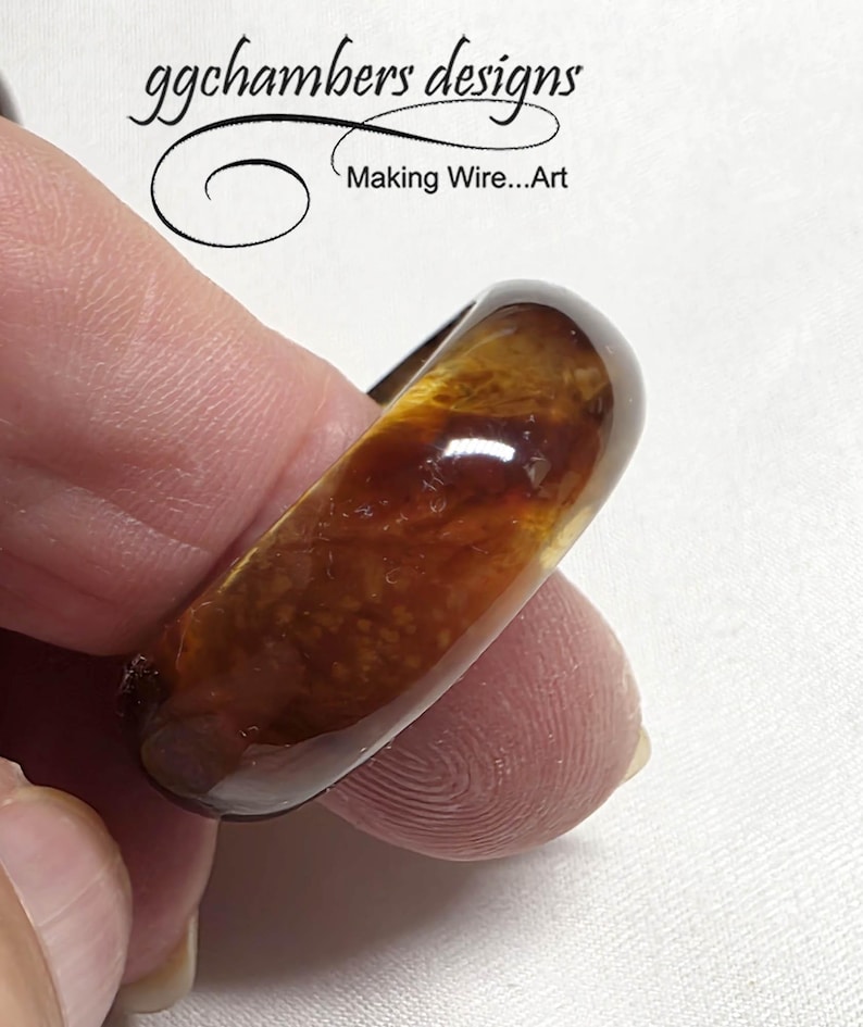 May include: An amber-coloured ring with a glossy finish. The ring has a unique, organic design with visible internal patterns. The text "ggchambers designs Making Wire...Art" is visible in the background.