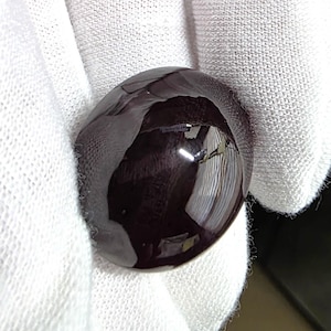 May include: A smooth, round, dark burgundy cabochon gemstone with a highly polished, reflective surface. The stone displays a deep, solid color and a domed shape, capturing light reflections on its glossy, curved top surface.