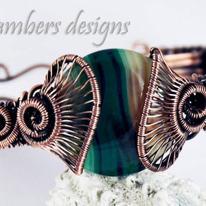 May include: A copper wire wrapped bracelet with a green and brown agate stone in the center. The wire is woven into a decorative pattern with swirls and fans.