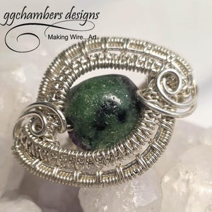 May include: A handcrafted silver wire pendant with a green and purple gemstone center. The pendant features intricate wire wrapping and the text "ggchambers designs Making Wire...Art". The gemstone is a deep green color.