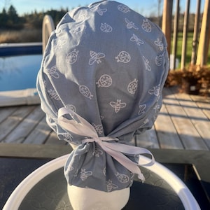 May include: A pale blue scrub cap featuring a white insect design of bees and ladybirds. The cap is tied with a white ribbon. The cap is displayed on a white form, set against a sunny outdoor backdrop.