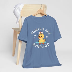 May include: A blue t-shirt featuring the text "Floated and Confused" and a cartoon duck wearing a nurse's cap. The duck is in water, with hearts around it. A white hat hangs on a wooden chair.