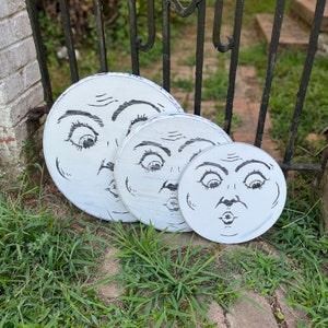 May include: Three white, round wooden plaques with a distressed finish, each featuring a whimsical, black-ink face of the moon. The sizes vary, creating a layered display. The plaques are arranged on a bed of green grass.