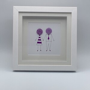 May include: A white frame with a white mount surrounding a white card with a purple and white button couple illustration. The couple are holding hands and the woman is wearing a striped dress and the man is wearing a tie.