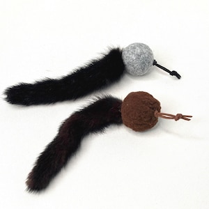 May include: Two cat toys, each featuring a long, dark brown fur tail attached to a round, textured ball. One ball is light gray felt with a black cord loop, and the other is dark brown textured fabric with a thin brown leather cord loop.