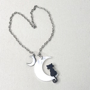 May include: A silver-toned metal chain necklace featuring two pendants. One pendant is a small, solid silver-colored crescent moon. The second, larger pendant is a white, pearlescent crescent moon with a small, black cat silhouette attached to it.